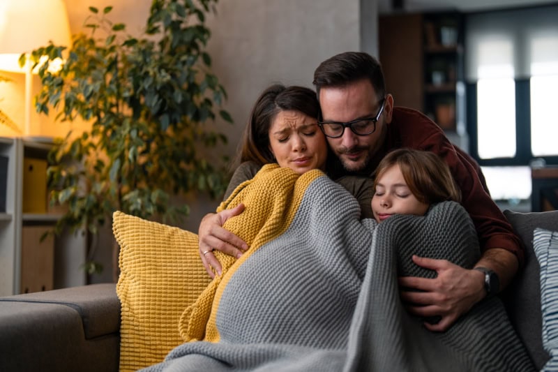 Why Isn’t My Furnace Heating My Entire Home? Photo of a family of three, cold and huddled together under a blanket trying to stay warm in their living room.